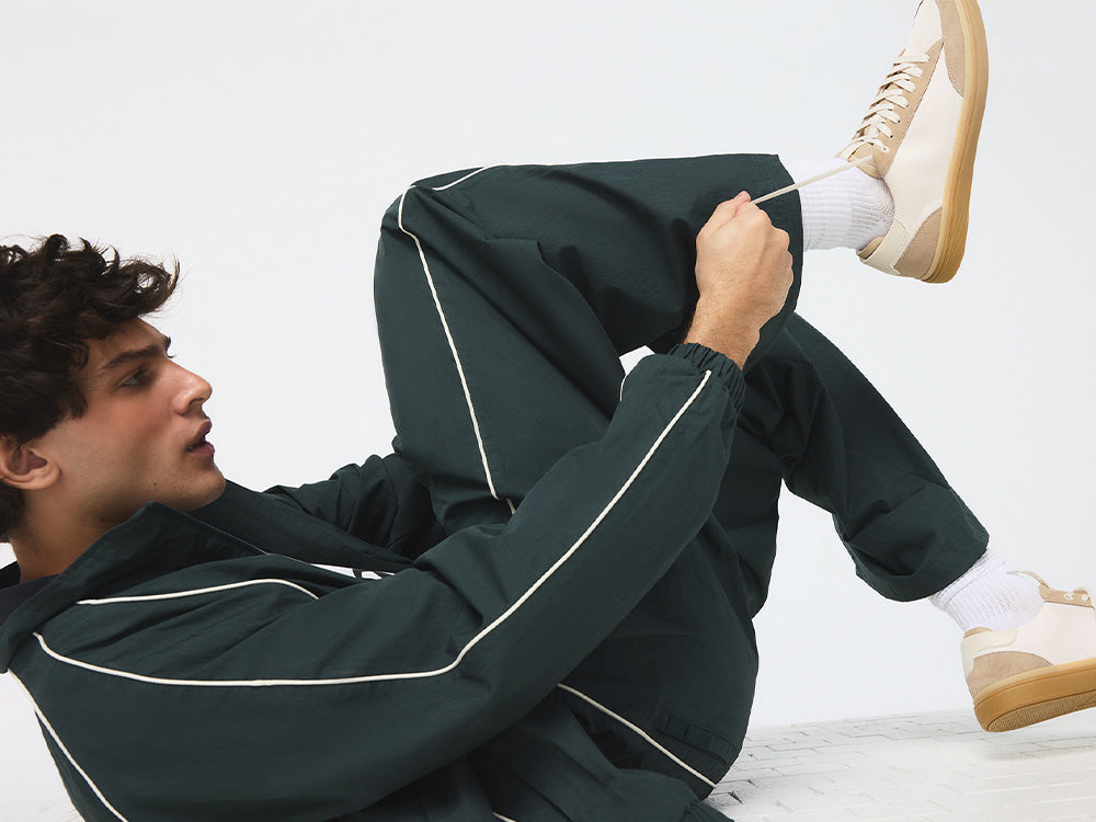 A man in a green tracksuit ties his shoelaces, wearing beige and white shoes.