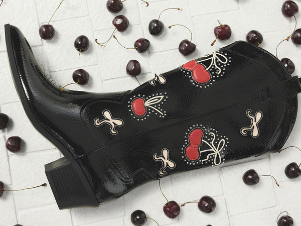 A black western boot with a cherry print is displayed against a white brick background with scattered cherries.